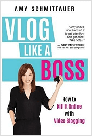 Amy Landino wrote the book Vlog Like a Boss under the name Amy Schmittauer. The cover shows a photo of Amy from the waist up holding a video camera. The title appears on a light blue background with white and fuchsia letters. The book's tagline is How To Kill It Online With Video Blogging.