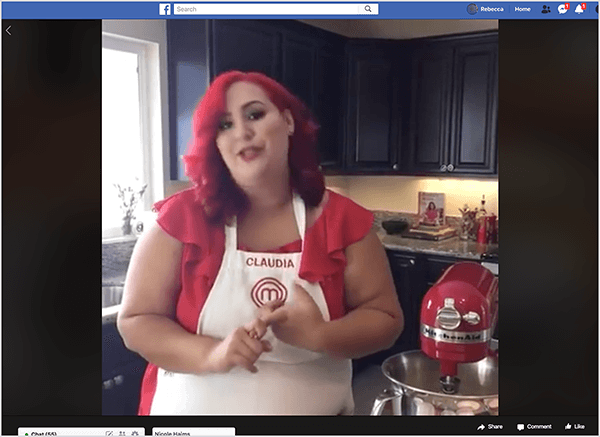 This is a screenshot of a Facebook Live video featuring Chef Claudia Sandoval, which she hosted in 2016 as part of a cross-promotion with the T-Mobile Tuesdays app. In the video, Claudia stands in a kitchen with black cabinets and granite countertops. A window over the sink lets natural light into the room. Claudia is standing next to a red Kitchen Aid mixer. She is a Latina woman who has bright red hair that falls just below her shoulders. She’s wearing makeup, a bright red top, and a white apron with Claudia and the MasterChef logo embroidered in red thread. As she’s talking, she’s gesturing with her hands. In 2016, Stephanie Liu collaborated with Claudia to produce this live video.