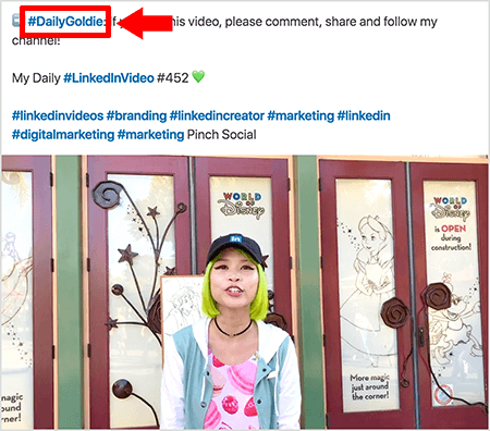 This is a screenshot that illustrates how Goldie Chan uses hashtags in the text of her LinkedIn video posts. Red callouts point to the #DailyGoldie hashtag in the text, which is unique to her video posts and helps her track shares. The post also includes other relevant hashtags that help people find her video, including #LinkedInVideo. In the video image, Goldie stands in front of some doors at a World of Disney display. She’s an Asian woman with green hair. She’s wearing a black LinkedIn cap, a black choker necklace, a pink macaron-print shirt, and a blue and white jacket.