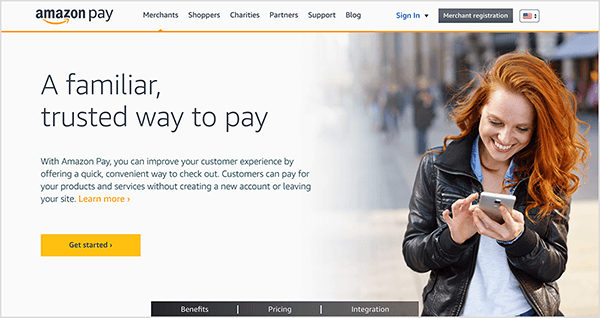 This is a screenshot of the Amazon Pay webpage. It has a white background and black text. The navigation options at the top are Merchants, Shoppers, Charities, Partners, Support, and Blog. A Sign In link and black Merchant Registration button also appear. In the main area of the web page, a heading and some other text appears on the right. The heading says “A familiar, trusted way to pay.” A yellow button labeled Get Started appears below the heading. On the right is a photo of a white woman with long red hair. She is standing and looking at her smartphone. She’s smiling and wearing and black jacked and button down shirt.